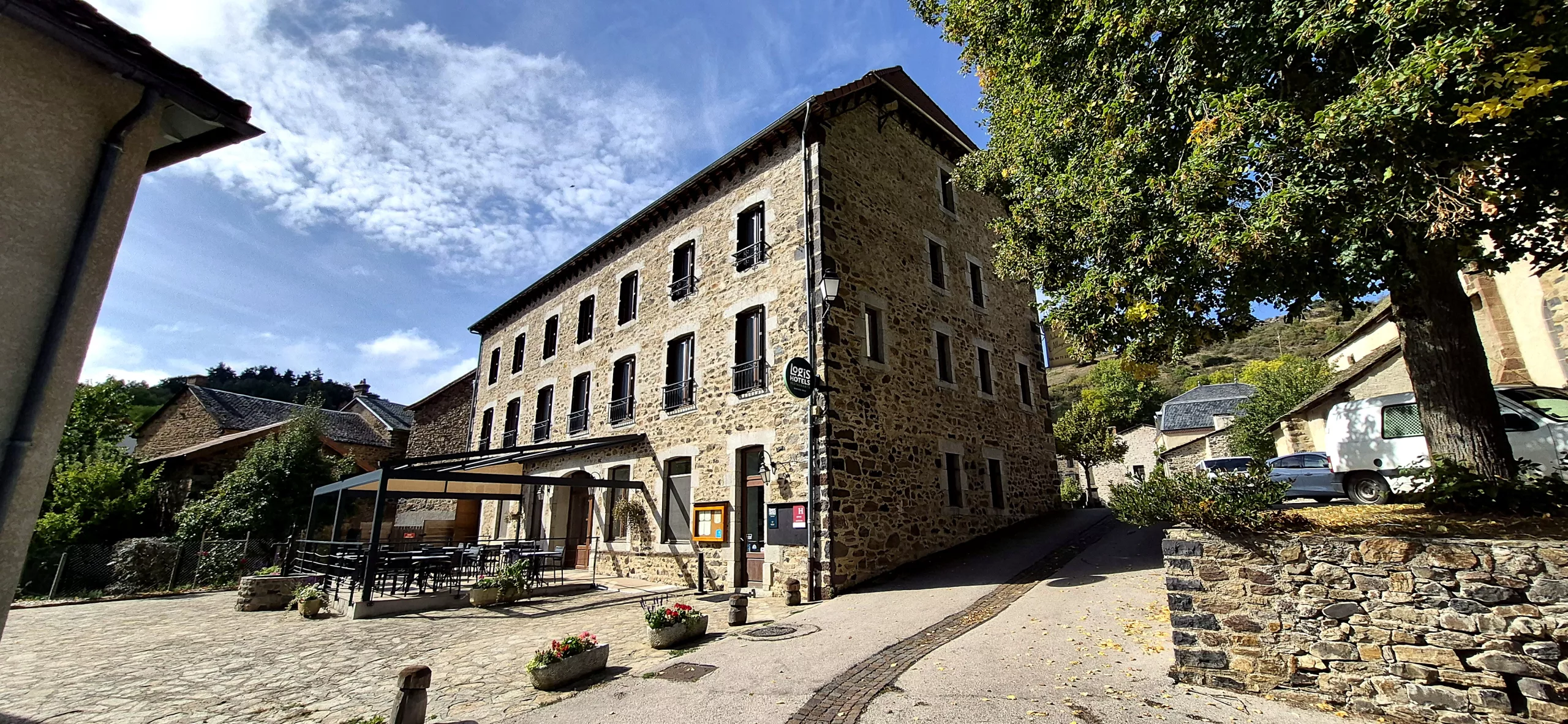 L'auberge en pierre à l'angle de la rue, sous un ciel bleu, avec sa terrasse et l'enseigne Logis Hôtels